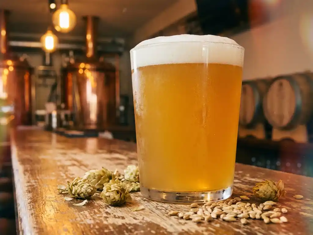 Goudkleurig IPA-bier met witte schuimkraag op rustieke houten bar, hop en gerst decoratief gerangschikt, brouwerijverlichting